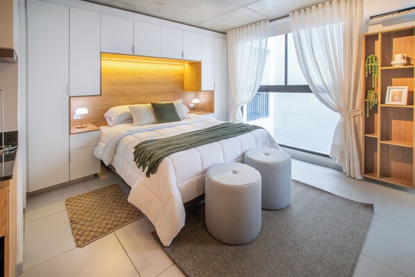 A modern bedroom with a bed, bedside lamps, a bedside table, a window with curtains, and a bookshelf. - Corporate Stays First del Sol - 604 (Asunción)