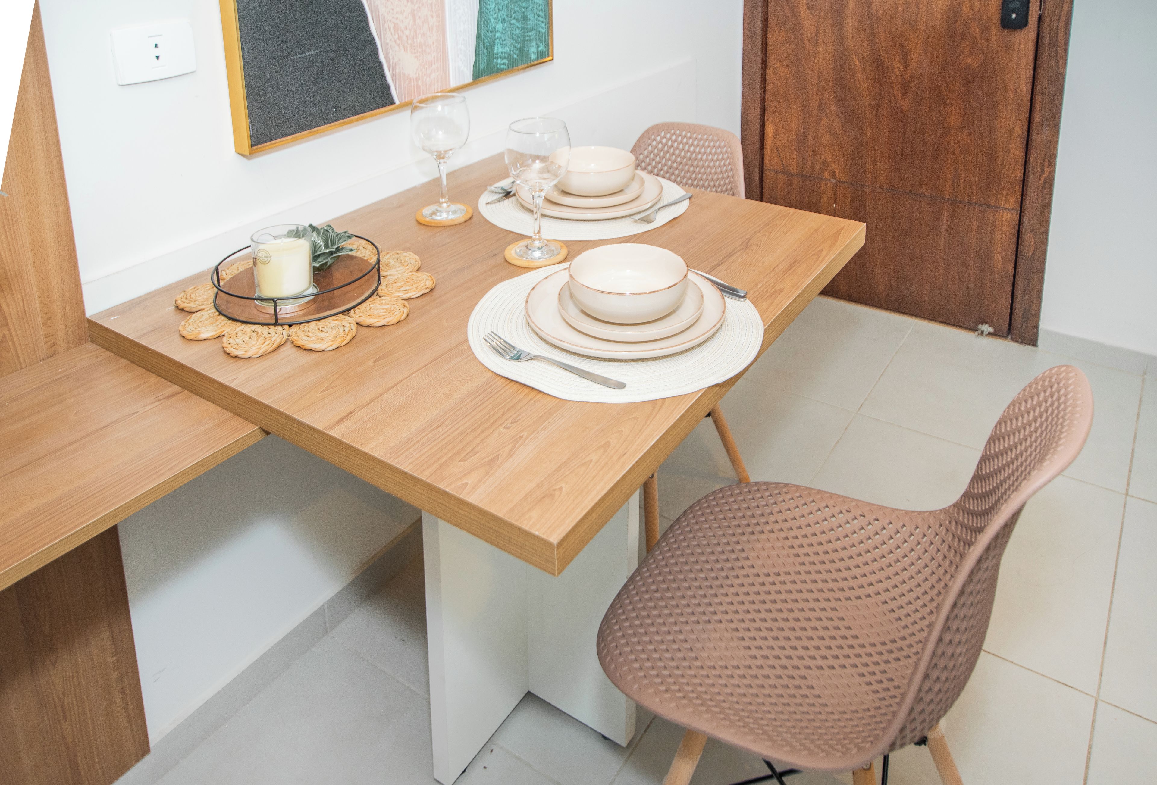 Una mesa de comedor dispuesta con platos, tazones, cubiertos y una vela, con una silla y un interruptor de luz en la pared.