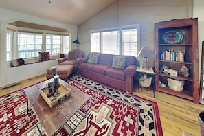 A living room with a sofa, a coffee table, a patterned rug, a bookshelf, and a window with blinds. - Gonzo Chalet | Spacious Deck + Mountain Views (Ruidoso)