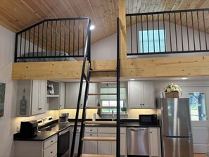 A kitchen with a wooden ceiling, a black staircase, white cabinets, a stainless steel refrigerator, and a window with a view. - Cozy Gruene Cottage - a short distance to Gruene and the Guadalupe River (New Braunfels)