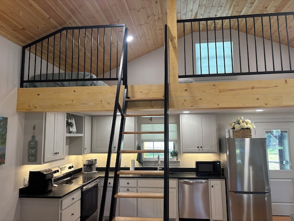 Una cucina con soffitto in legno, una scala nera, mobili bianchi, un frigorifero in acciaio inossidabile e una finestra con vista.