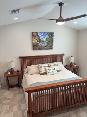 A bedroom with a wooden bed, a ceiling fan, a nightstand with a lamp, and a wall-mounted painting of a river. - Cozy Gruene Cottage - a short distance to Gruene and the Guadalupe River (New Braunfels)
