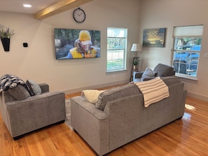 A living room with two grey sofas, a large flat-screen TV, a clock, and a window with a view of parked cars. - Cozy Gruene Cottage - a short distance to Gruene and the Guadalupe River (New Braunfels)