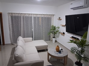 Ein Wohnzimmer mit einem Ecksofa, einem Couchtisch, einem Flachbildfernseher und zwei Topfpflanzen.