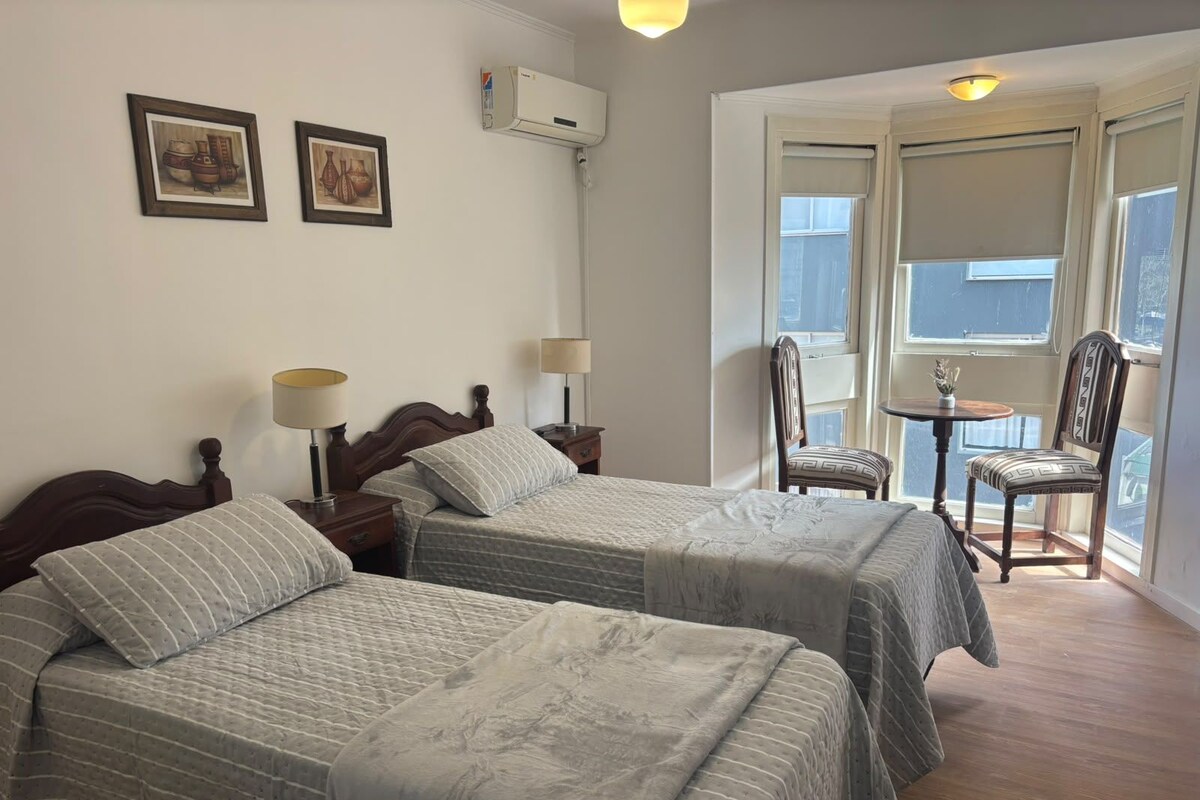 A hotel room with two beds, a small table, chairs, and a window with a view.