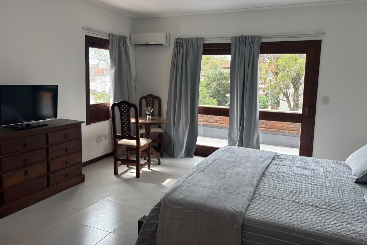 A hotel room with a bed, a TV on a dresser, a dining table with chairs, and a balcony with curtains.
