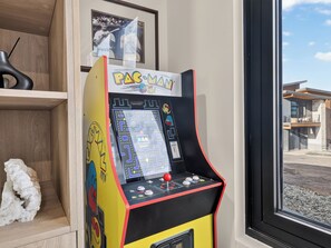 A Pac-Man arcade machine in a room with a window, a framed photo on the wall, and a small white sculpture on the floor. - Ski-In/Out Luxury | Private Hot Tub + Sauna (Brian Head)