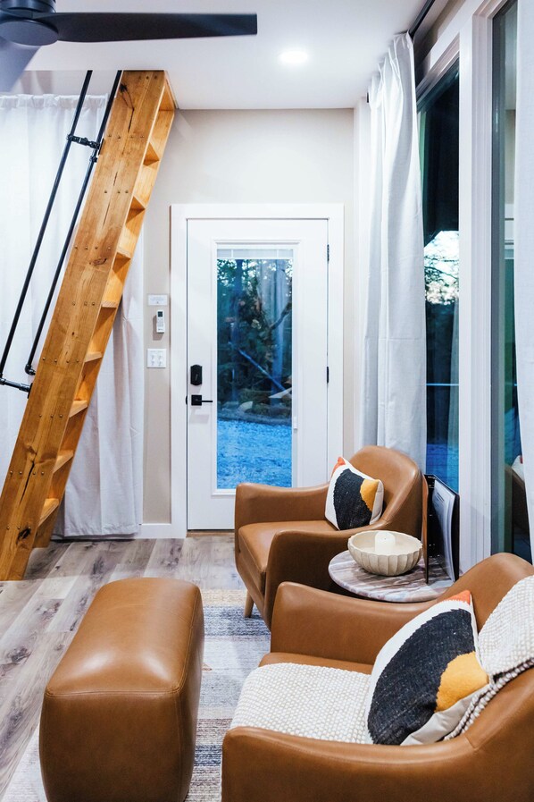 A modern living room with a wooden staircase, leather sofas, and a view of a blue outdoor area. - Hemlock | Pond Views + Hot Tub, Game Room & Kayaks (Campton)
