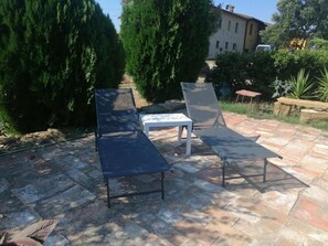 A patio with two lounge chairs and a small table, surrounded by stone tiles and greenery.