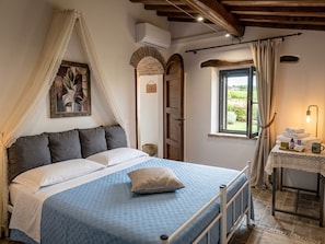 A bedroom with a bed, a bedside table with a lamp, a window with curtains, and a wall-mounted air conditioner.