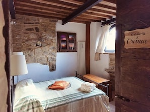 A rustic bedroom with a stone wall, wooden beams, a bed with a green and white bedspread, a bedside table, and a window with a curtain. - Vecchio Casale Quercetino  (Montenero)