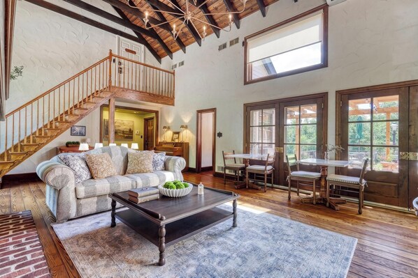 A spacious living room with a wooden staircase, a large window, and a dining area with wooden furniture.