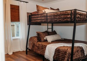 Un lit superposé avec une couverture matelassée marron et des oreillers, un plaid blanc et une fenêtre avec des rideaux blancs.