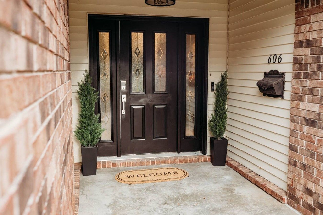 A dark wooden front door with diamond patterns, a welcome mat, and two potted plants on either side.