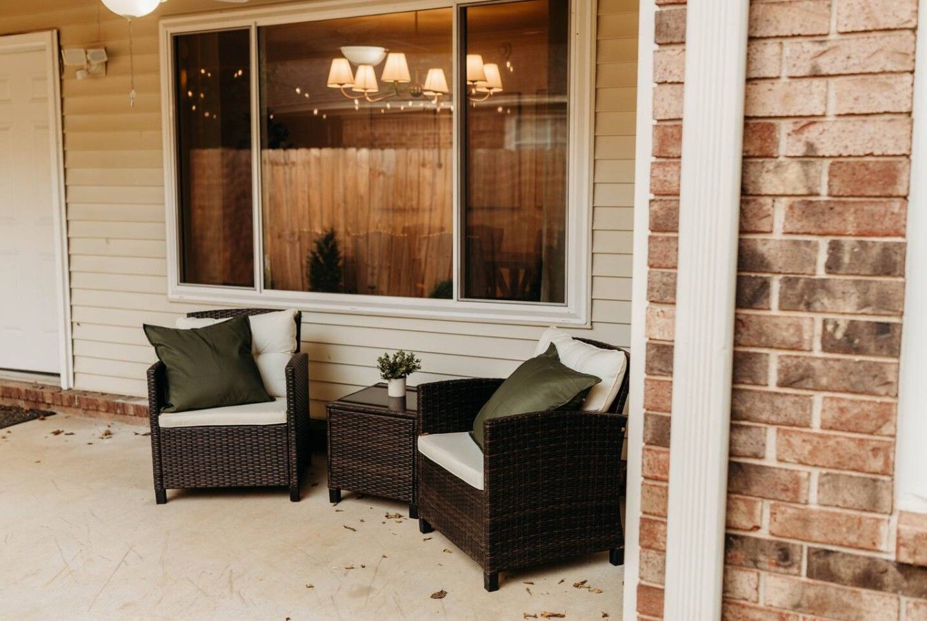 A covered porch with two wicker chairs and a small table, a window with curtains, and a brick wall.