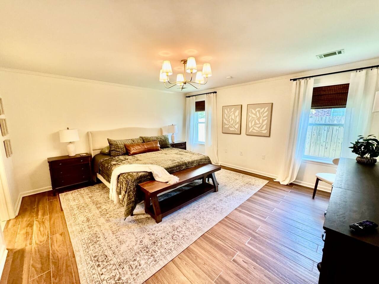 Een slaapkamer met een bed, nachtkastjes, een bankje, een kroonluchter en een raam met gordijnen.