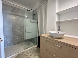 Een badkamer met een douchecabine van glas, een witte wastafel en een houten opbergmeubel.