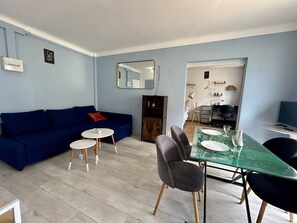Ein Wohnzimmer mit einem blauen Sofa, zwei kleinen Tischen, einem Glastisch mit Stühlen und einer Küche im Hintergrund.