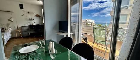 A dining area with a table set for two, a television, and a balcony with a view of the sea.