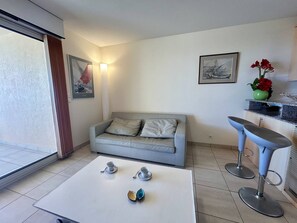 Ein modernes Wohnzimmer mit einem grauen Sofa, einem weißen Couchtisch mit Teekannen, einer Küchenzeile mit Barhocker und Kunstwerken an den Wänden.