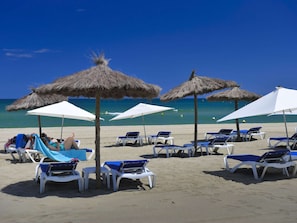 Ein Strand mit weißen Liegestühlen und Strohdächern, einige davon von Personen genutzt.