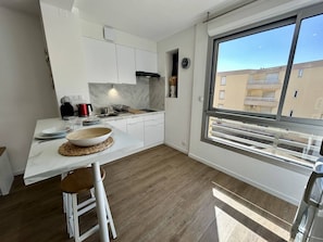 Ett modernt kök med vita skåp, en väggbeklädnad av marmor, ett stort fönster med utsikt över byggnader och en matplats med en skål av trä och ätpinnar.