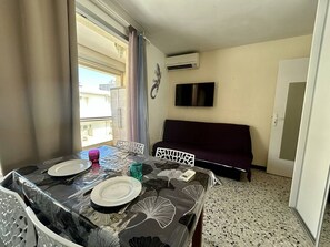 A dining area with a table set for two, a sofa, a TV, and an air conditioning unit. - Studio with AC near beach - 4 guests (Canet-en-Roussillon)