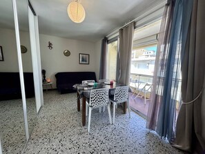 A room with a dining table, chairs, a sofa, a small side table, and a window with curtains. - Studio with AC near beach - 4 guests (Canet-en-Roussillon)