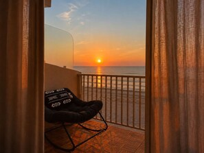 Ein Balkon mit Blick auf den Sonnenuntergang über dem Meer, ein schwarzer Stuhl mit gemustertem Kissen und ein Holzboden.