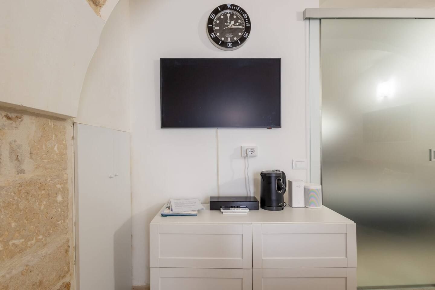 Een kamer met een wandgemonteerde flatscreen televisie, een koffiezetapparaat, een afstandsbediening en een wit kastje.