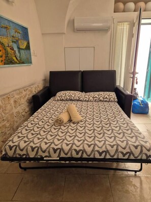 Ein Bett mit einem gemusterten Bettüberwurf, ein Paar zusammengerollte Handtücher, ein schwarzer Kopfteil und ein Gemälde an der Wand.
