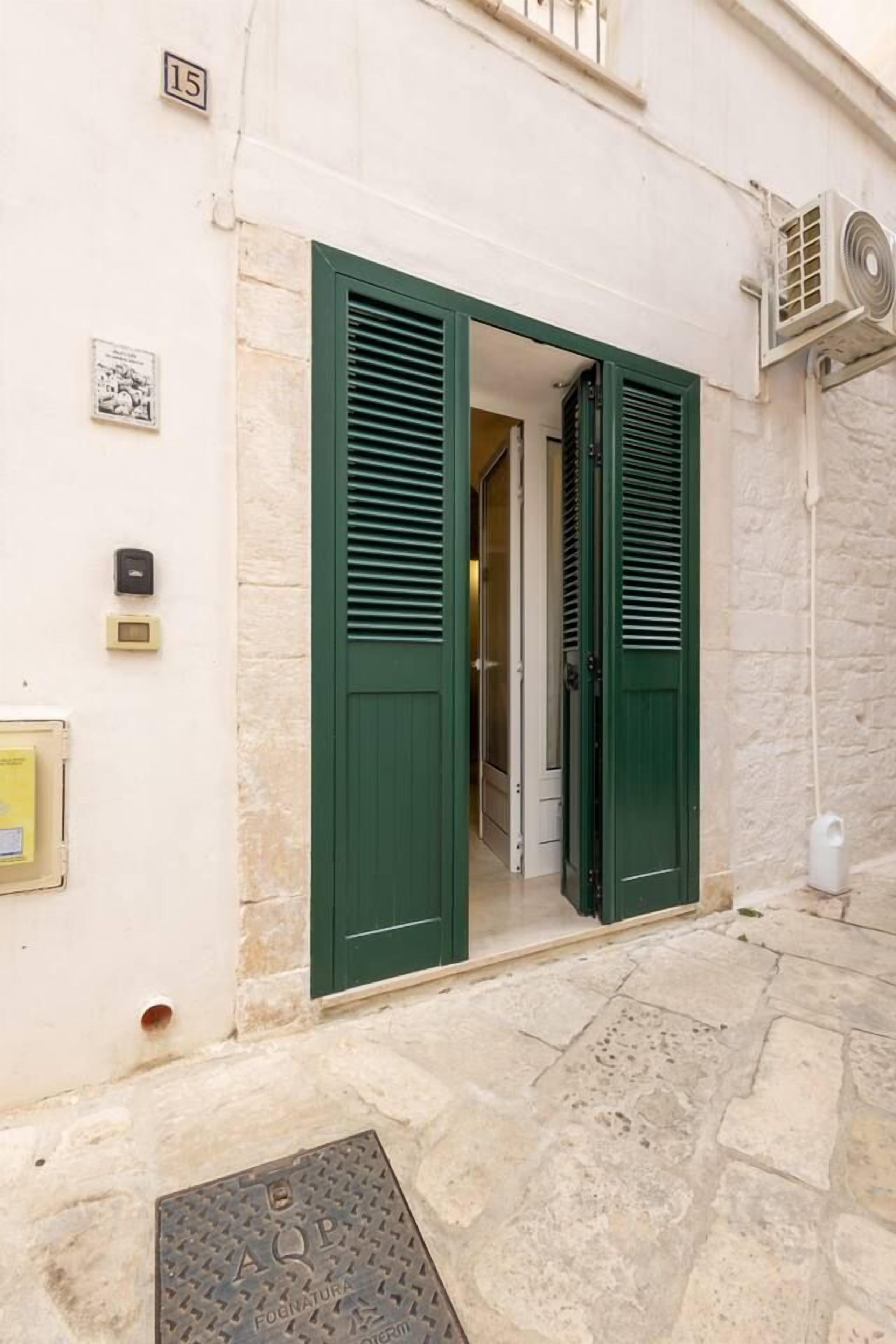 Une entrée d’immeuble avec des volets verts, une plaque portant le numéro « 15 », et un petit espace extérieur doté d’un sol en pierre.