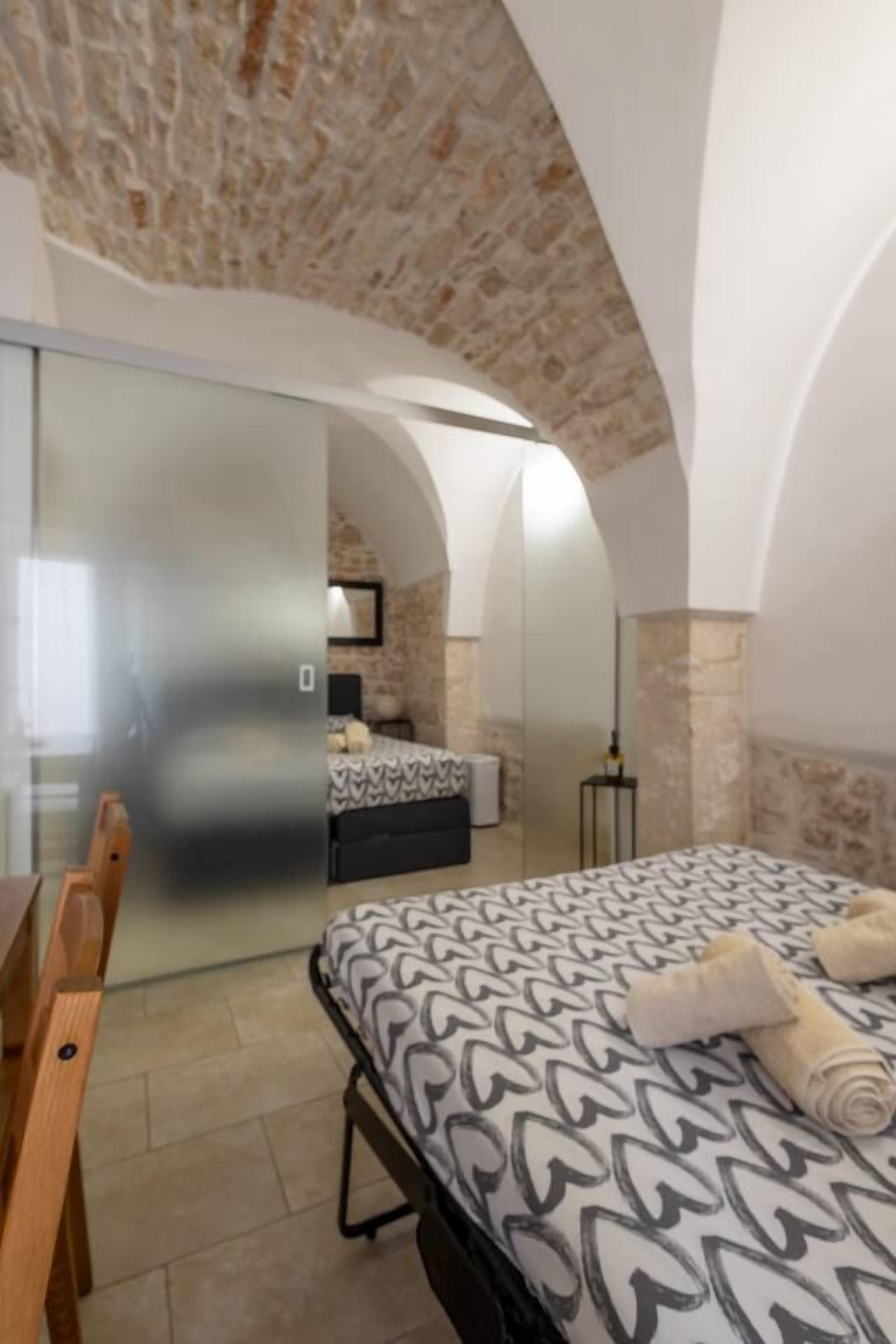 Ein Schlafzimmer mit einem Bett, einem Schreibtisch und einem Stuhl. An der Wand hängt ein Spiegel und es gibt ein Fenster.