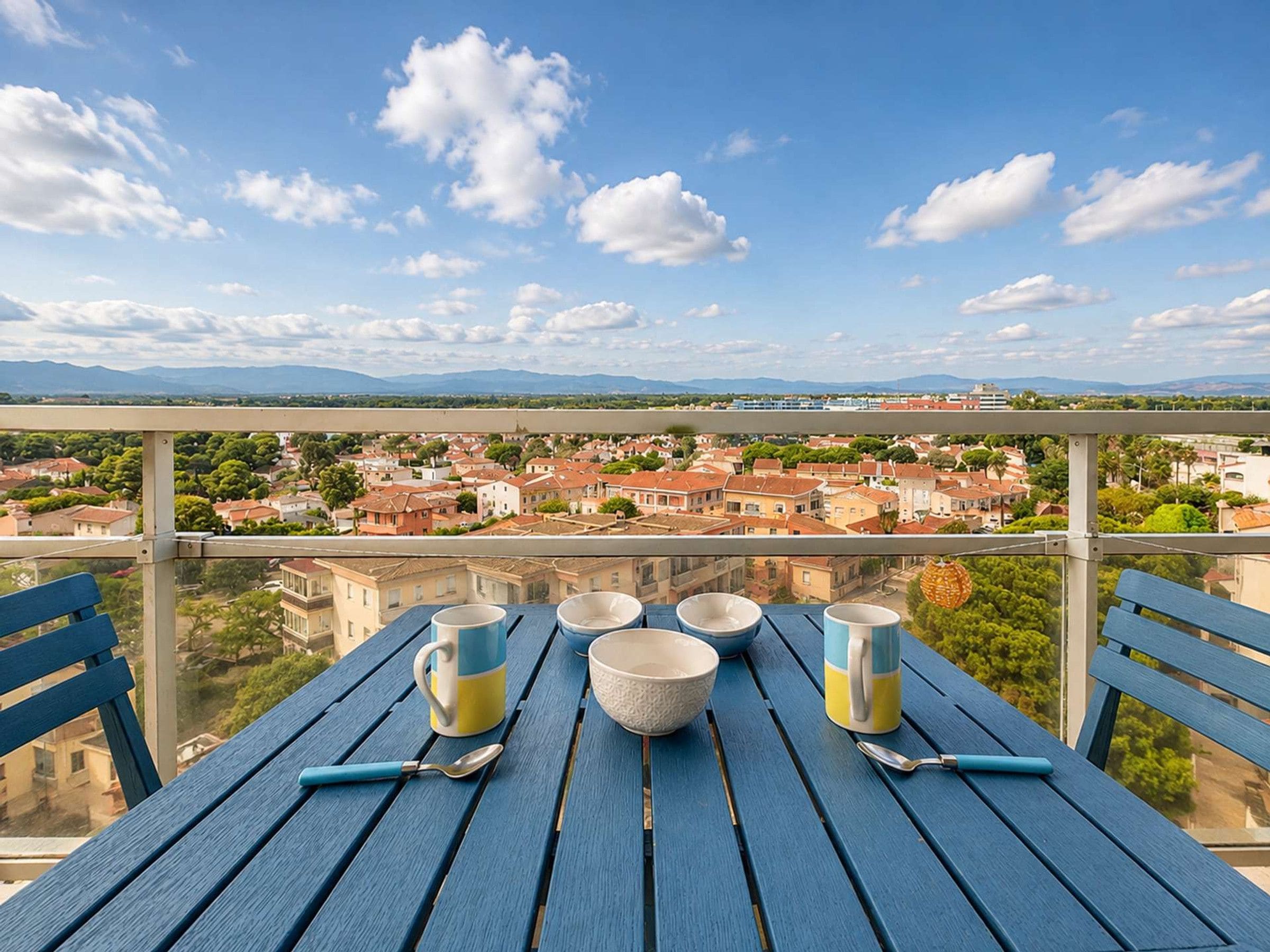 A balcony with a table set for dining, overlooking a town with buildings and mountains in the distance.