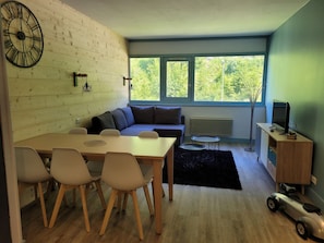 Ein stilvoller Wohnraum mit Essbereich, einer Couch, einem Fernseher und einem großen Fenster mit Blick auf Grünflächen.