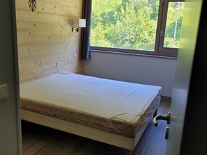 A room with a wooden wall, a bed with a mattress, a window with a view of trees, and a door with a handle. - Spacious apartment near slopes - 8 guests (Flaine)