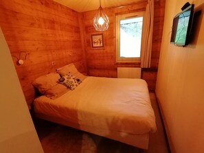 Un dormitorio de cabaña de madera con cama, lámpara colgante, ventana con cortinas y televisor montado en la pared.