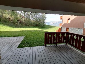 Una terraza de madera con barandilla, un césped y un edificio de paredes de piedra al fondo.