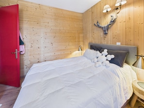 A bedroom with a wooden wall, a red door, a bed with white bedding, and a wall-mounted lamp. - Village center apartment, balcony - 4 guests (Les Gets)