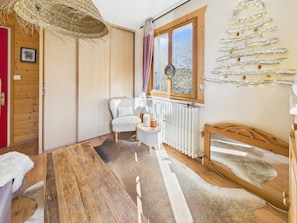 A cozy room with a wooden floor, a white chair, a wooden bed, a radiator, and a window with a view of trees. - Village center apartment, balcony - 4 guests (Les Gets)