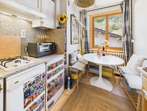 A compact kitchen with a gas stove, a microwave, and a dishwasher. There is a round dining table with a white tablecloth and two chairs. A window with curtains is visible, and a painting hangs on the wall. - Village center apartment, balcony - 4 guests (Les Gets)