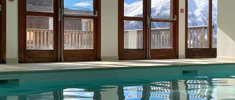 Uma piscina com reflexos nítidos, amplas janelas com molduras de madeira e vista para montanhas cobertas de neve.