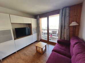 A living room with a flat-screen TV, wooden cabinets, a purple sofa, a small wooden table, and a sliding glass door leading to an outdoor balcony. - Apartment - 6 guests, mountain view (Orcières Merlette)