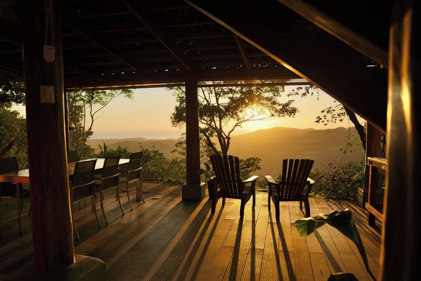 Terraza de madera con sillas y una mesa, con vistas a un paisaje montañoso al atardecer.