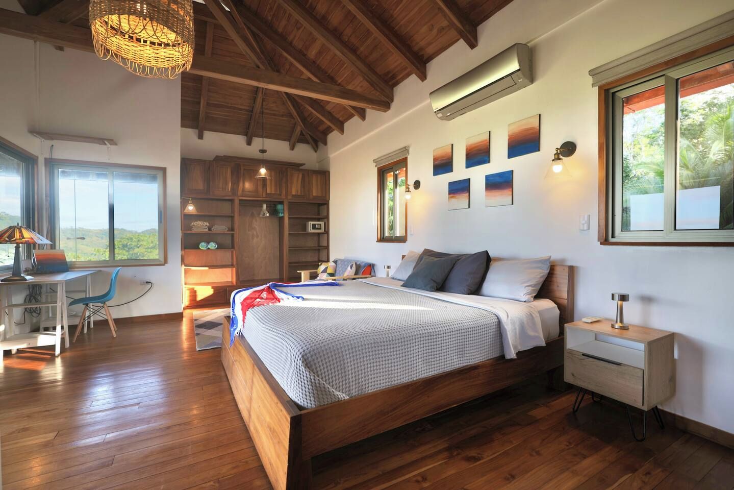 A bedroom with a wooden bed, bedside tables, a desk, and a chair. There are windows with a view of greenery outside.