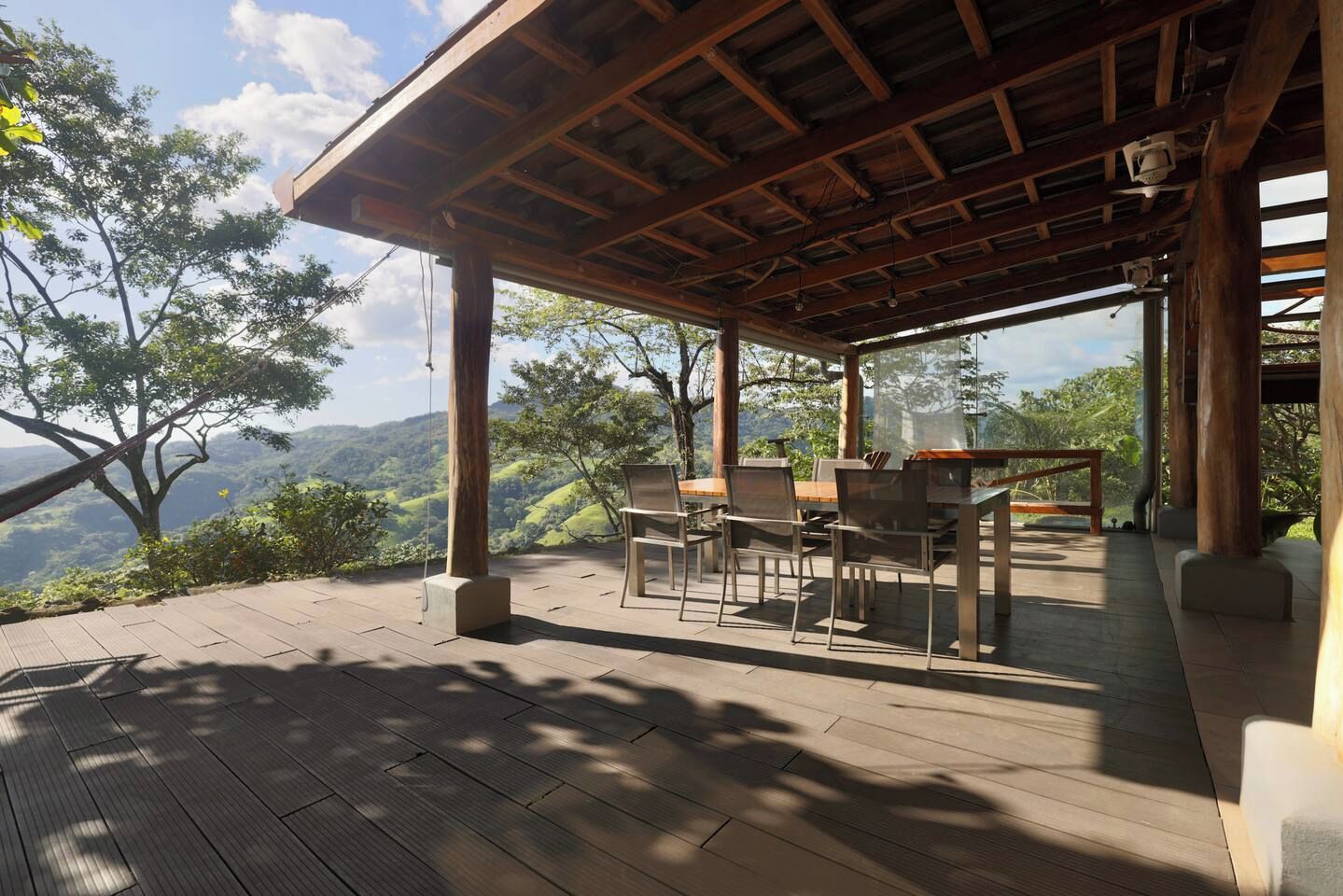 Een terras buiten met een houten tafel en stoelen, omgeven door een houten terras en uitzicht op een bergachtig landschap.