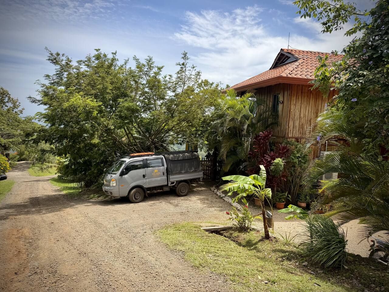 Een vrachtwagen geparkeerd voor een huis met een rode pannendak, omgeven door groen en een grindpad.