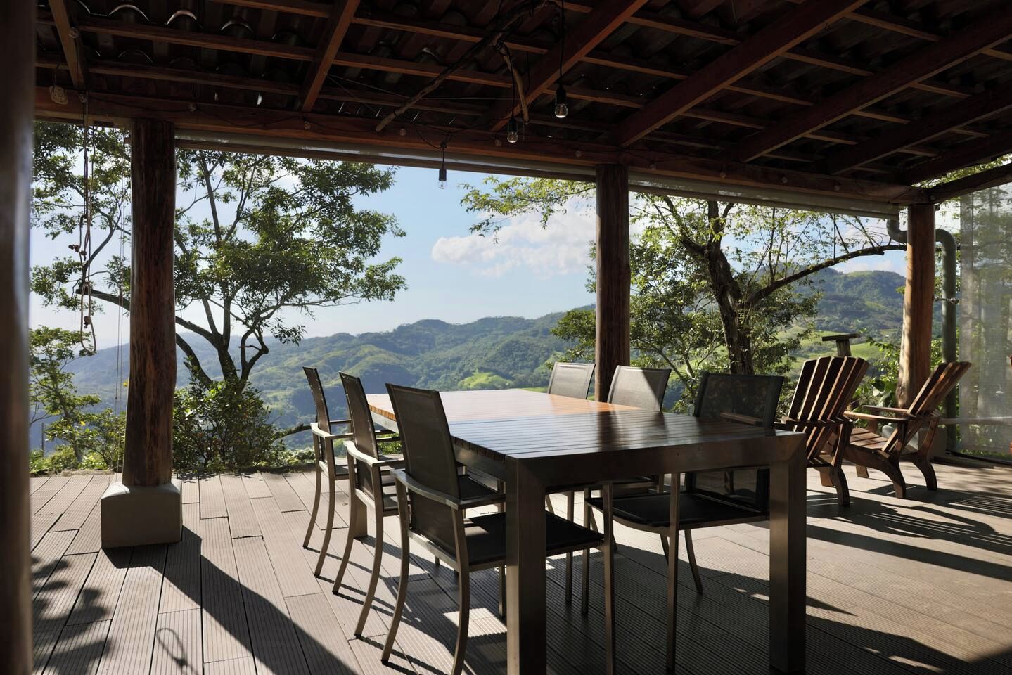 An outdoor dining area with a table and chairs, surrounded by wooden pillars and a view of a mountainous landscape.