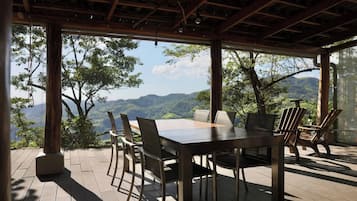 An outdoor dining area with a table and chairs, surrounded by wooden pillars and a view of a mountainous landscape.