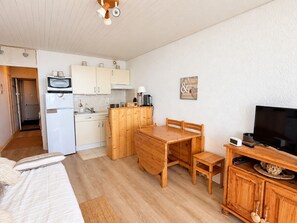 A compact kitchen with a microwave, stove, and cabinets, a wooden dining table with chairs, a television on a wooden stand, and a bed with a white comforter. - Apartment, panoramic view - 4/5 guests (Chamrousse)